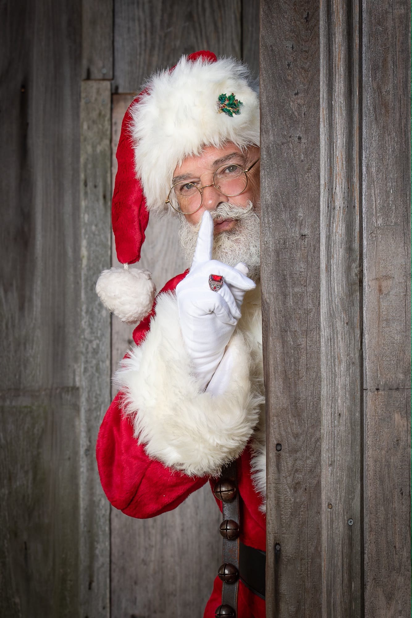 Santa Joe in a traditional red suit peeking around a wooden door with a finger to his lips